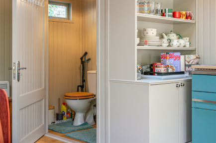 Toilet installed inside hipped garden room View from the kitchen area into a compact bathroom, with built-in shelving and cabinetry beside the doorway.